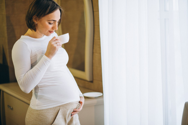 young pregnant woman standing by window drinkig coffee 1303 21076
